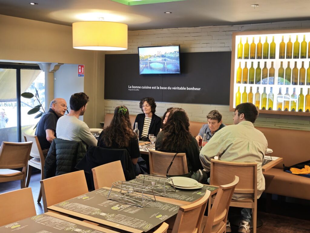 Large group seated around a long table at a Parisian restaurant, wine bottles displayed on the wall behind them