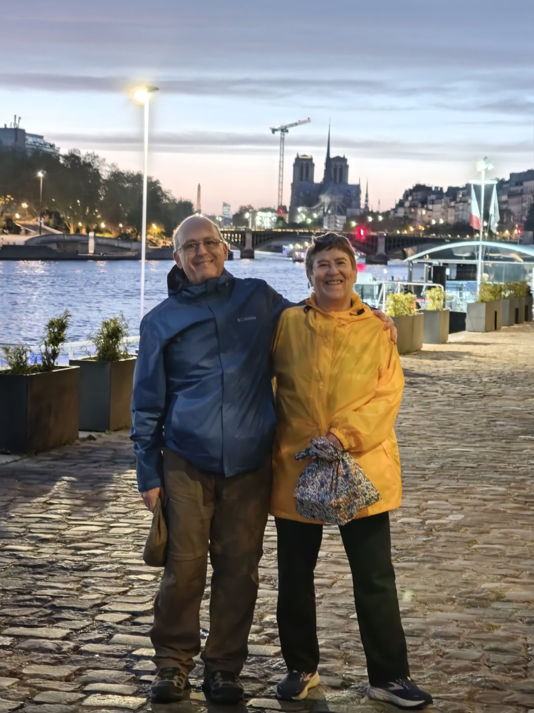 Ben and Kathy standing on a cobblestone quayside at night with Notre-Dame cathedral illuminated across the Seine