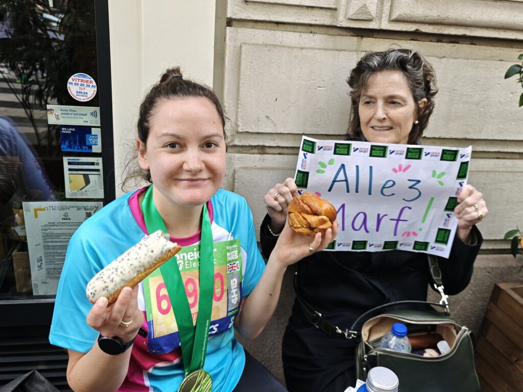 A woman in marathon gear holding an eclair while another holds a handmade Allez Marf sign