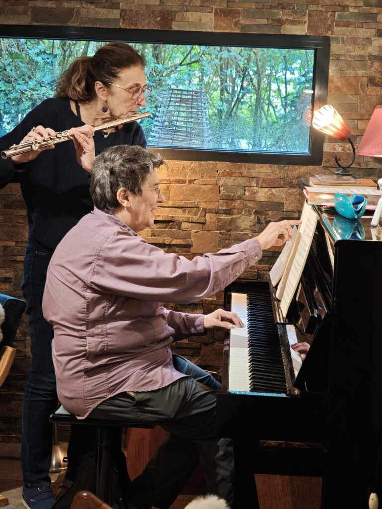 Two women playing flute and piano in a cozy stone-walled room with warm lamp light