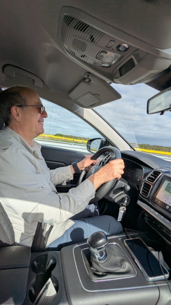A man smiling at the wheel on a French highway with yellow rapeseed fields visible through the window