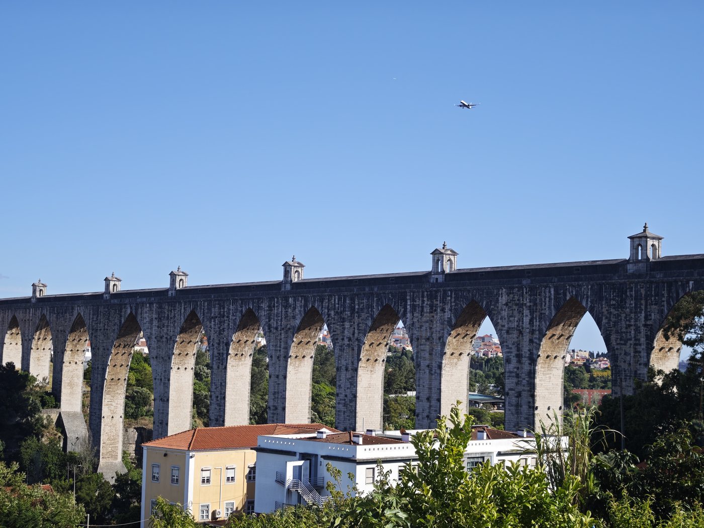 The Águas Livres Aqueduct with a plane passing overhead against a blue sky
